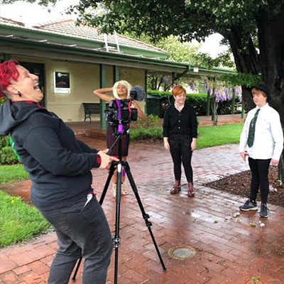 Someone laughs behind a camera as four young people smile in front of it under a tree