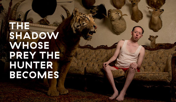 A man sits on a vintage couch in a singlet an shorts next to a taxidermy tiger
