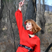 Woman with a red bob haircut wearing a bright red dress strikes a dance pose with right arm extended in the air