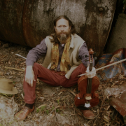 A man in red pants and a grey shirt with long brown hair and beard crouches in front of a tree