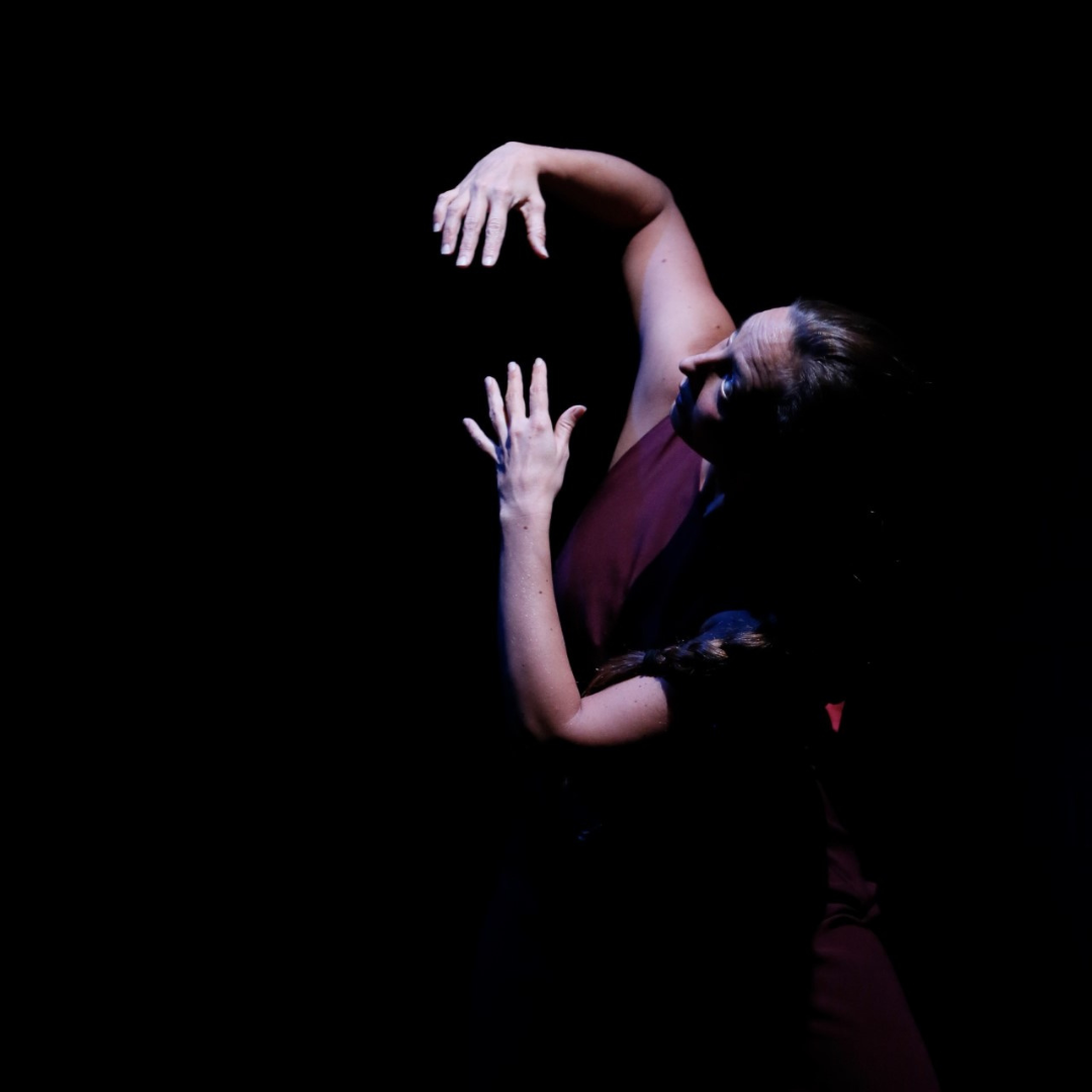 A woman in stage lighting striking a dance posed with one hand reaching up and wone hand reaching down to meet it