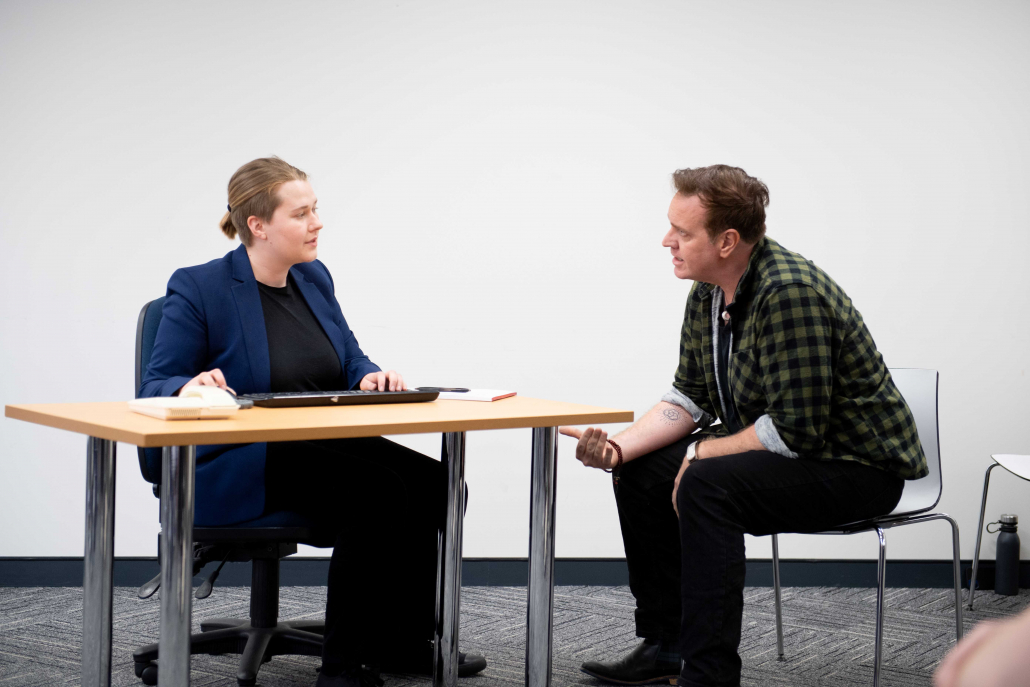 two people face each other across a desk, one with blue suit jacket and undercut hair tied back in a bun, the other wearing a green checkered shirt with short brown hair