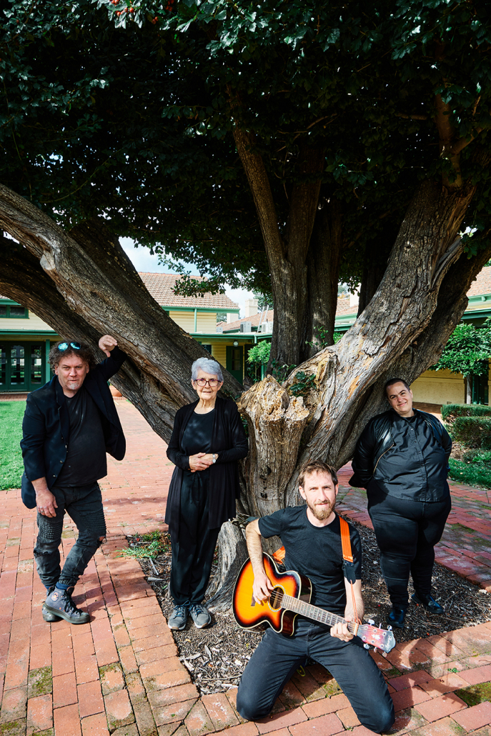 4 people in black clothes sit/stand below a big tree posing for the camera
