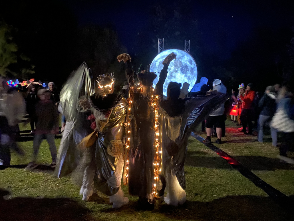 Four Young Women in Moth Costumes stand in dance poses with a big blue moon sculpture in the background