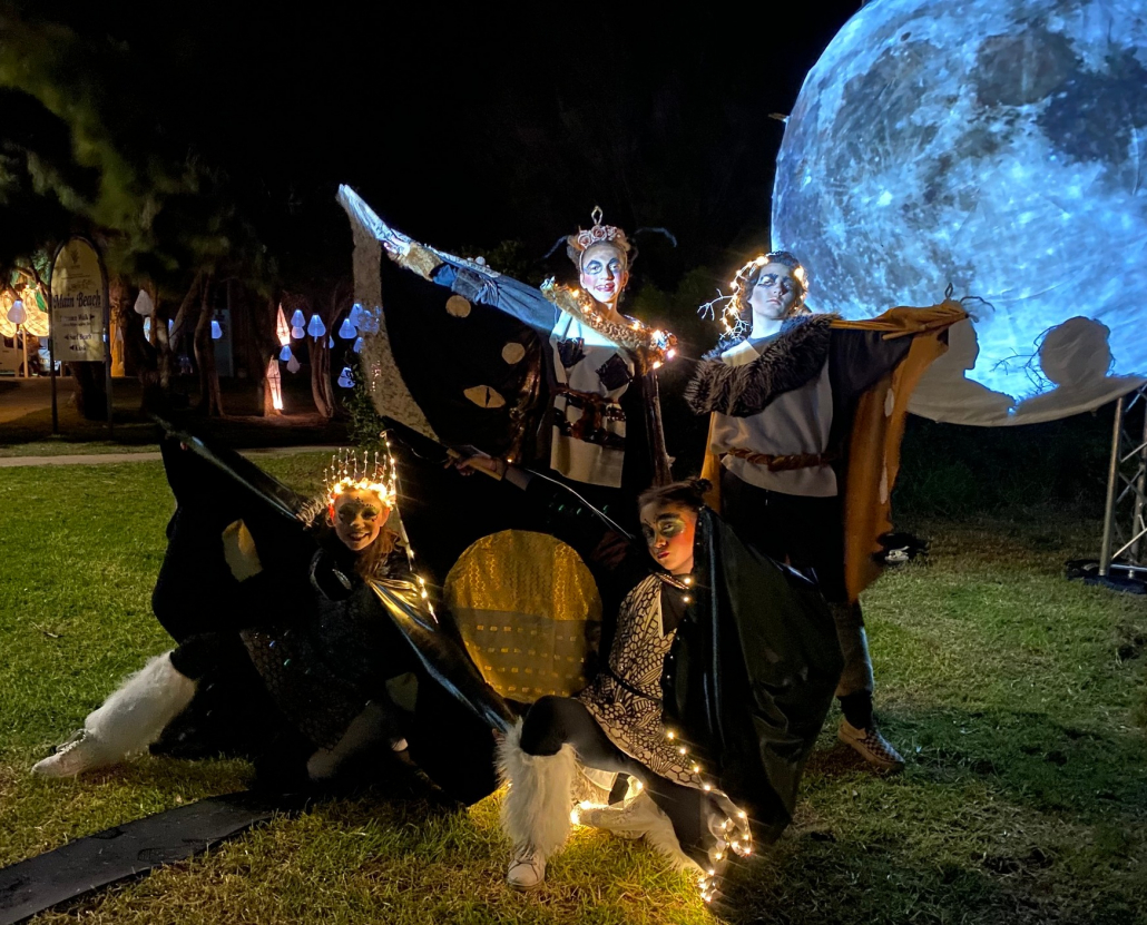 Four Young Women in Moth Costumes stand in dance poses with a big blue moon sculpture in the background