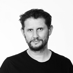 A man in a black tee shirt with short beard and short brown hair smiles at camera