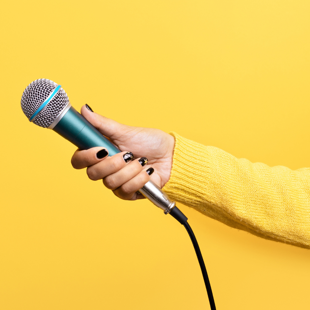 A hand is extended holding a microphone on a yellow background. The arm holding the microphone is covered a yellow woollen sweater