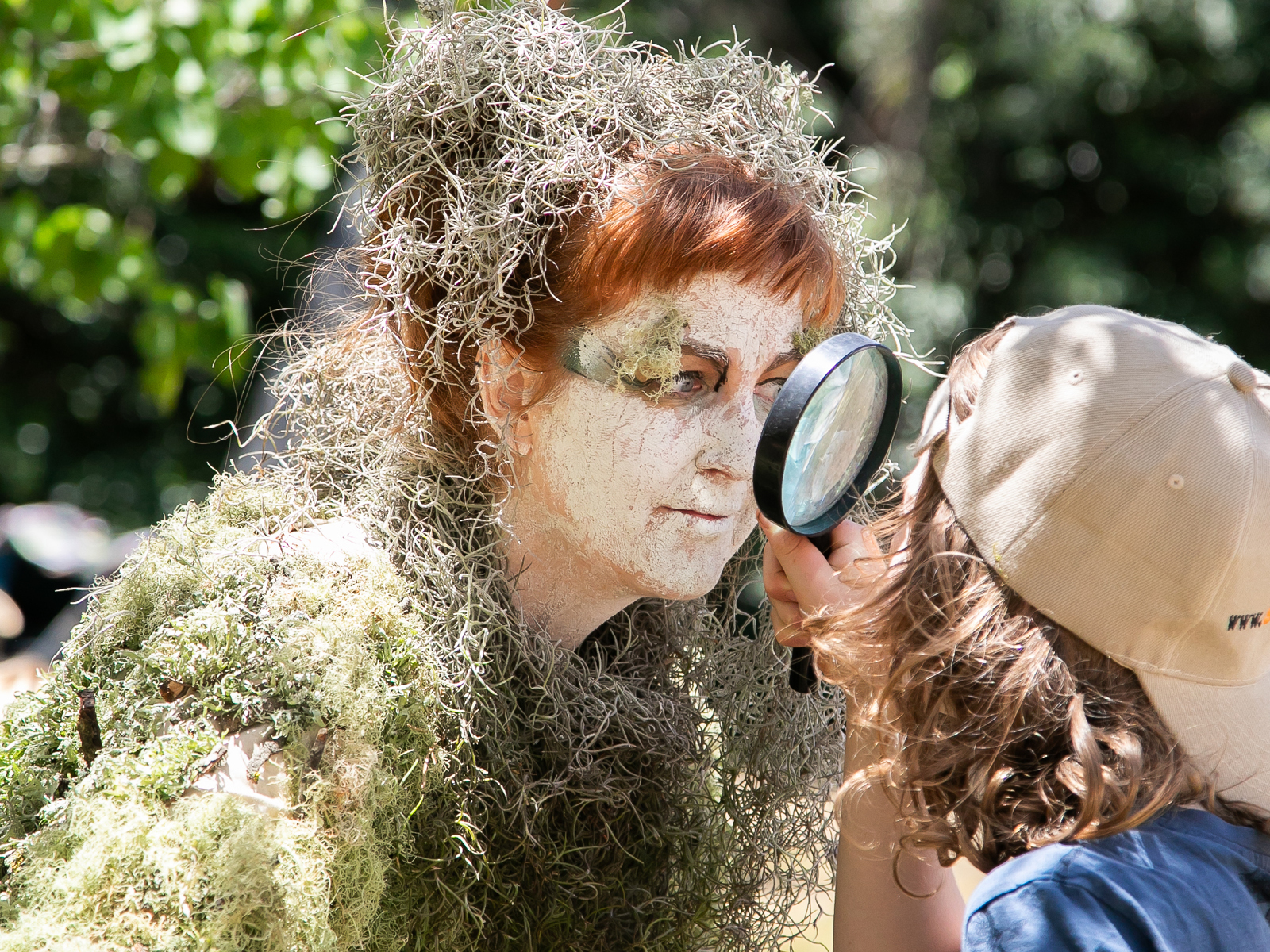 A Woman in a costume made of moss stares at a child through a magnifying glass.