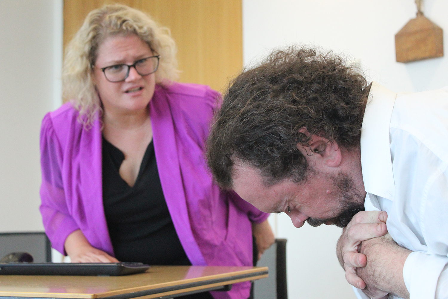 A Woman in a pink Jacket sits at a desk on the looks on as a man is buckled over in front of her