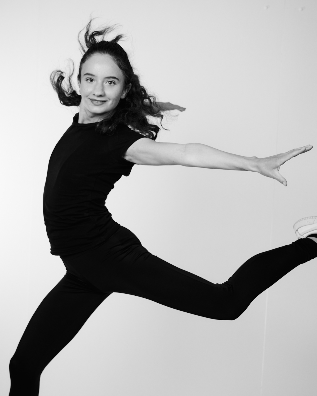 A woman in a black shirt smiling at the camera while doing a dancing jump wiht arms spread out to the sides