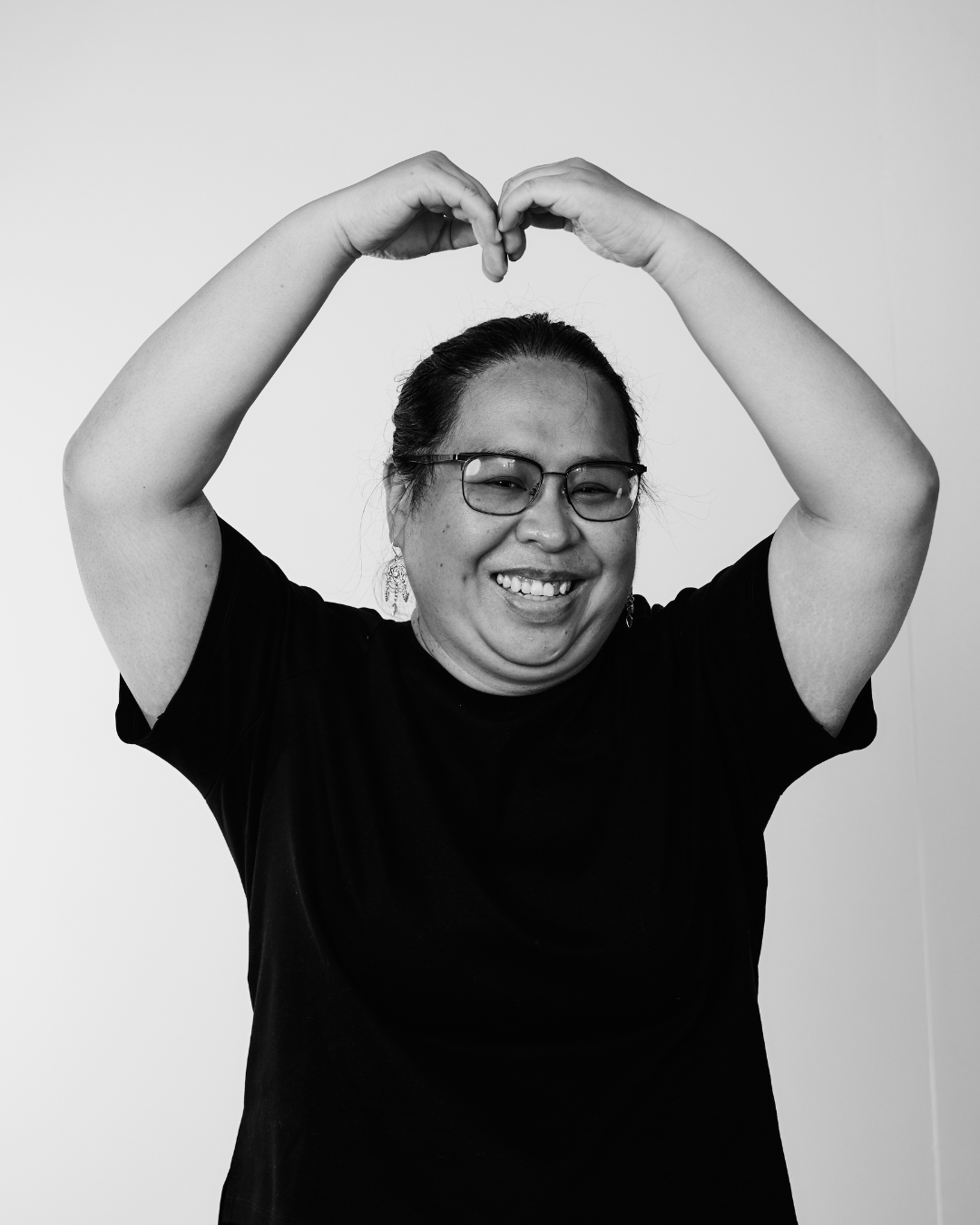 A woman in a black shirt smiling at the camera creating a love heart above hear head with her arms