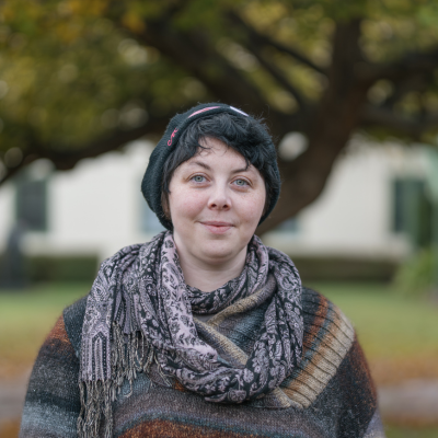 A persin in a grey and black sweater and scarf smiles at the camera on front of a large tree