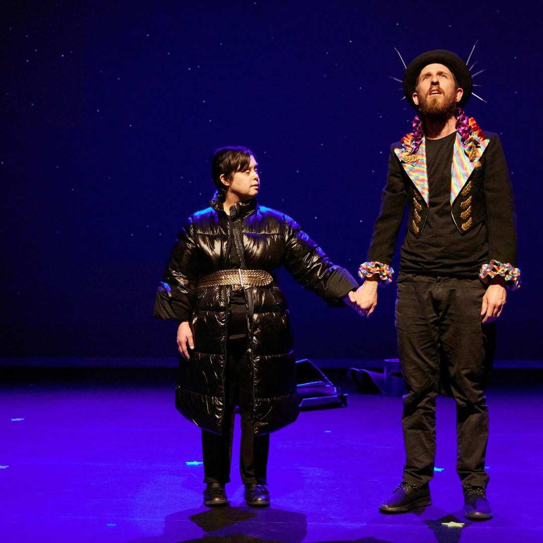 A man and a woman stand on stage in dramatic lighting